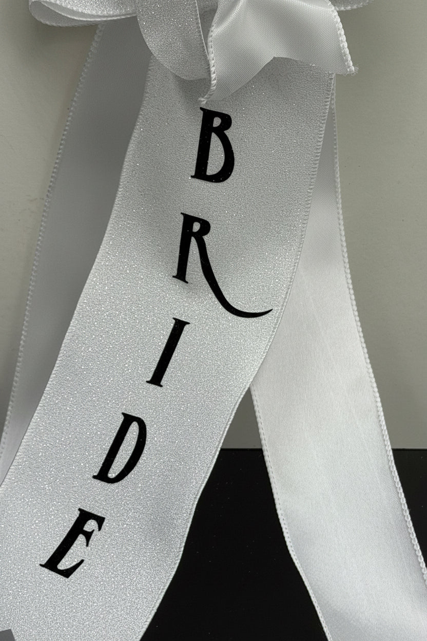 A white glitter ribbon sash printed with the word 'BRIDE' in black serif letters, shown against a neutral background.