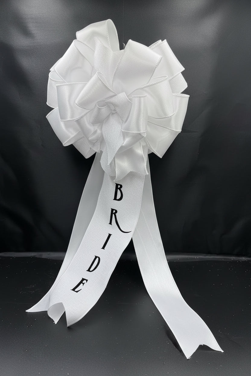 A white satin bridal bouquet bow with long tails printed with the word 'BRIDE' in black, displayed against a white backdrop inside a black box.