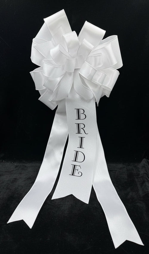 A white glitter and satin bridal bow with a long ribbon tail printed with the word 'BRIDE' in black capital letters, displayed on a white stand against a black background.