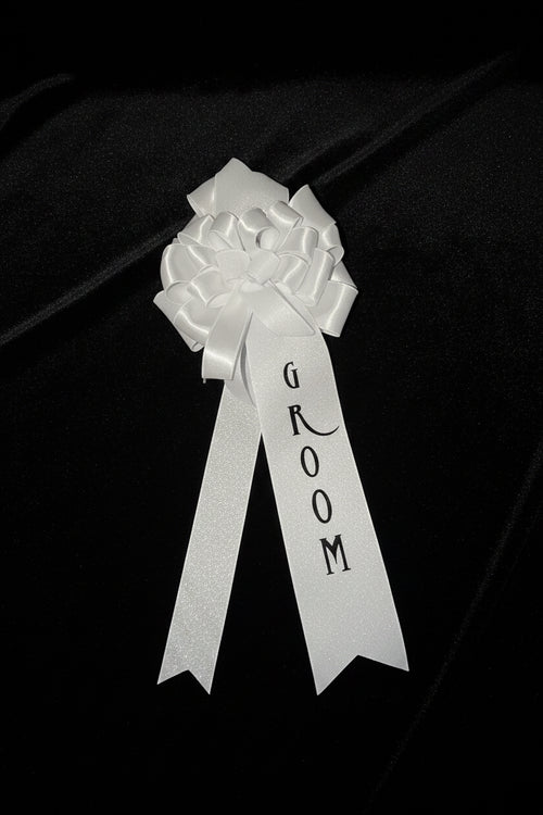 A white satin and glitter groom ribbon with the word 'GROOM' printed in black, attached to a white glitter and satin bow, photographed inside a black box.