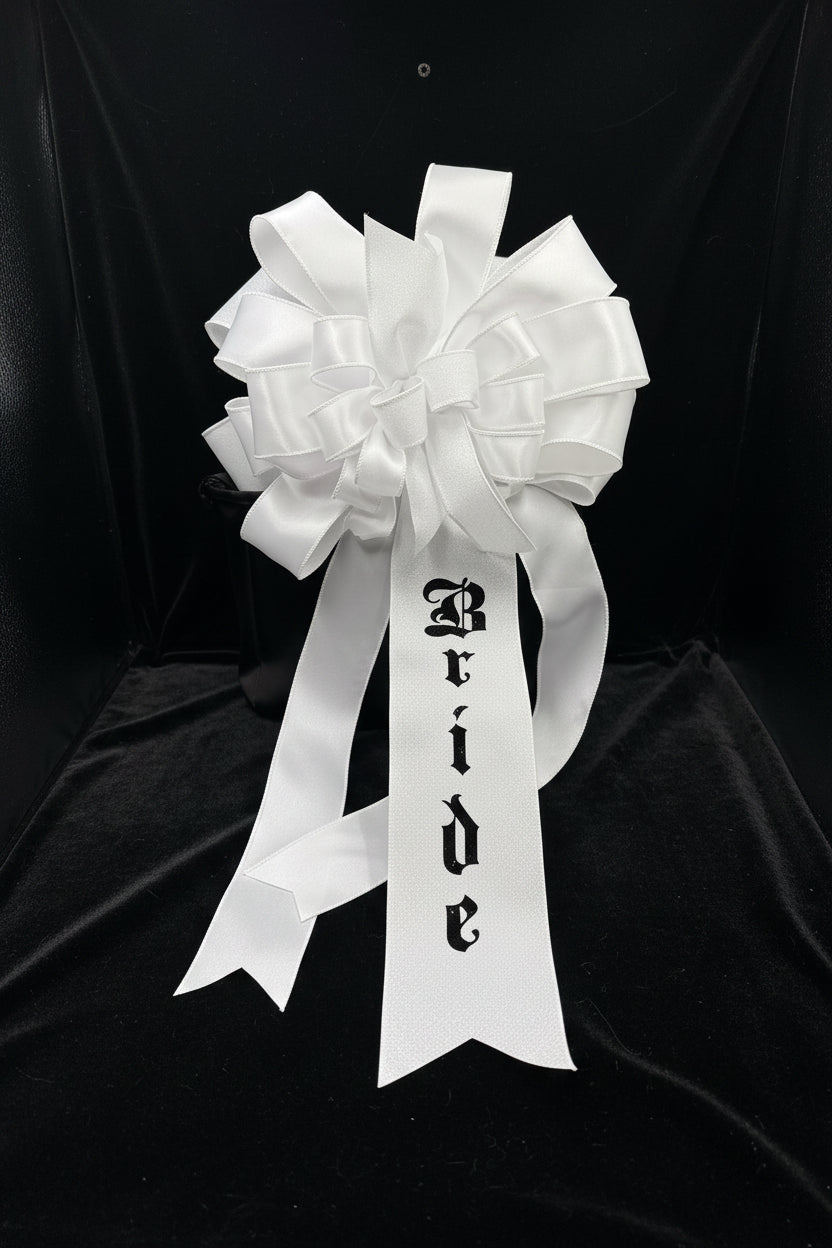 A white glitter and satin bridal bow with a long ribbon tail printed with the word 'Bride' in a black gothic font, displayed against a black background.