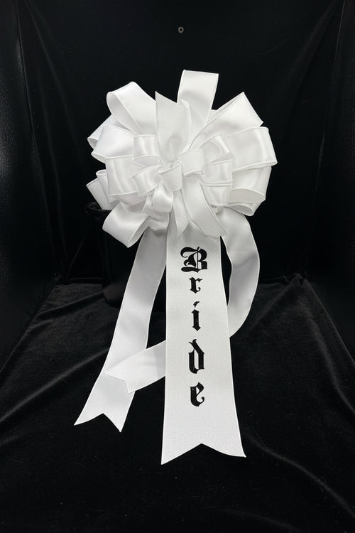 A white glitter and satin bridal bow with a long ribbon tail printed with the word 'Bride' in a black gothic font, displayed against a black background.